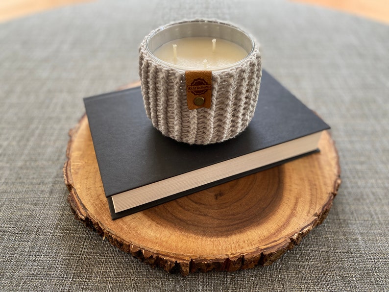 K&ouml;nnte beinhalten: Ein grauer gestrickter Kerzenhalter mit einem braunen Leder-Etikett mit der Aufschrift "Handmade with Love" steht auf einem schwarzen Buch, das auf einer Holzscheibe liegt.