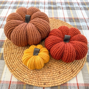 May include: Three knitted pumpkins in shades of orange and yellow sit on a round, woven placemat. The pumpkins have black leather-like stems. The background is a plaid fabric.