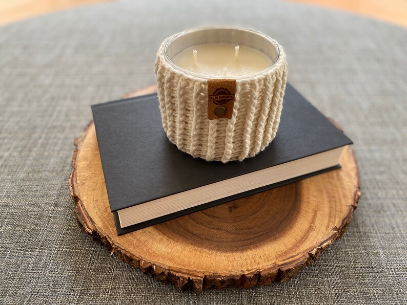 K&ouml;nnte beinhalten: Ein wei&szlig;er geh&auml;kelter Kerzenhalter mit einem braunen Leder-Etikett mit der Aufschrift "Wild Spirit" auf einem schwarzen Buch und einer Holzscheibe.