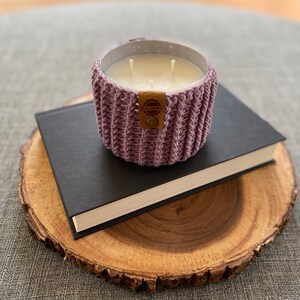K&ouml;nnte beinhalten: Ein lila geh&auml;kelter Kerzenhalter mit einem braunen Leder-Etikett mit der Aufschrift "Handcrafted" auf einem schwarzen Buch und einer Holzscheibe.