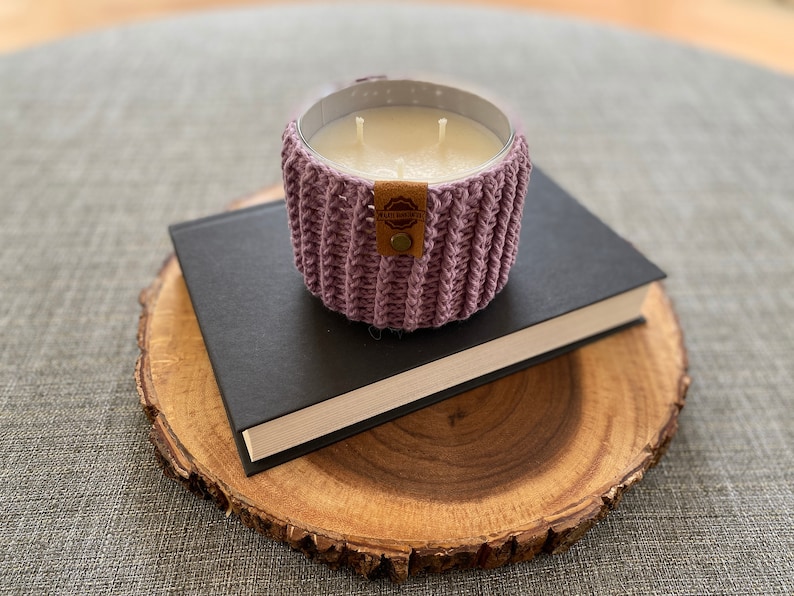 K&ouml;nnte beinhalten: Ein lila geh&auml;kelter Kerzenhalter mit einem braunen Leder-Etikett mit der Aufschrift "Handcrafted" auf einem schwarzen Buch und einer Holzscheibe.