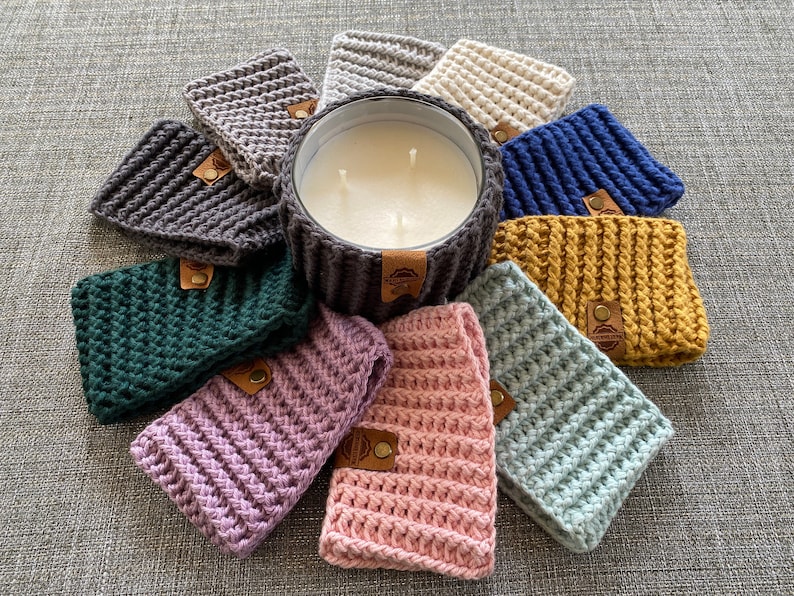 K&ouml;nnte beinhalten: Eine Sammlung geh&auml;kelter Kerzenw&auml;rmer in verschiedenen Farben, darunter Gr&uuml;n, Blau, Rosa, Lila und Gelb. Die W&auml;rmer sind um eine wei&szlig;e Kerze in einem Glasgef&auml;&szlig; angeordnet.