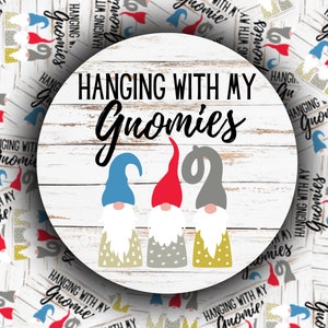 May include: A round wooden sign with a whitewashed background and the text "Hanging with my Gnomies" in black script. Three gnomes are depicted, one with a red hat, one with a blue hat, and one with a gray hat. Each gnome has a white beard and a patterned body.