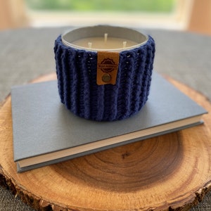 K&ouml;nnte beinhalten: Ein blauer geh&auml;kelter Kerzenhalter mit einem braunen Leder-Etikett, auf dem "Made Honestly" steht. Der Kerzenhalter steht auf einem grauen Buch und einer Holzscheibe.