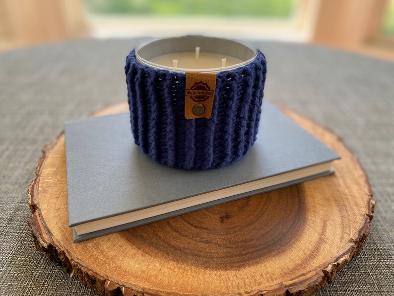K&ouml;nnte beinhalten: Ein blauer geh&auml;kelter Kerzenhalter mit einem braunen Leder-Etikett, auf dem "Made Honestly" steht. Der Kerzenhalter steht auf einem grauen Buch und einer Holzscheibe.