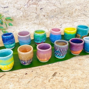 May include: A collection of small, hand-painted ceramic cups in various colors, including blue, green, yellow, and purple. The cups are arranged on a green leaf, with white flowers and foliage in the background. Each cup has a unique design.