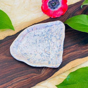 May include: A small, white ceramic dish with a speckled, blue and pink glaze. The dish is shaped like a triangle with rounded edges and is sitting on a wooden surface.