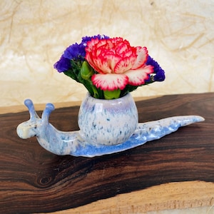 May include: A whimsical ceramic snail vase with a blue and white speckled design, holding a vibrant arrangement of red, white, and purple flowers. The snail is placed on a dark wooden surface, creating a charming decorative piece.