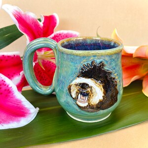 May include: A green ceramic mug with a brown and white dog face design. The mug has a handle and is sitting on a green leaf.