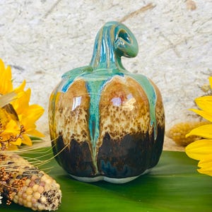 May include: A ceramic pumpkin sculpture featuring a glossy glaze with a color gradient transitioning from a dark brown base to a speckled tan middle and a vibrant teal top, complete with a curved, sculpted stem extending from the center.