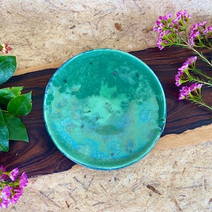 May include: A green ceramic plate with a mottled glaze. The plate is round and has a slight lip. It is sitting on a wooden surface.