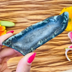 May include: A dark blue and green ceramic dish with a rough, textured surface. The dish is shaped like a long, narrow boat.