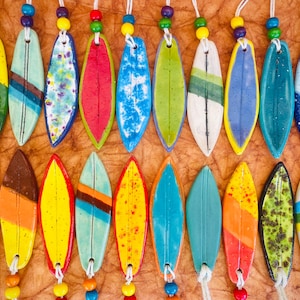 May include: A collection of 20 colourful ceramic surfboard ornaments with various patterns and designs. Each ornament has a string for hanging.