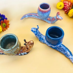 May include: Three ceramic snail-shaped planters in shades of blue, brown, and tan. Each planter has a round opening for plants. The snails are arranged on a light yellow surface, with colorful flowers in the background.