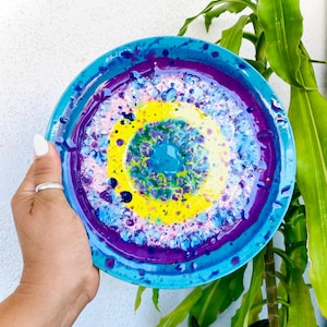 May include: A round decorative plate featuring a vibrant abstract design with concentric rings of bright turquoise, deep purple, and sunny yellow. The surface displays a textured, marbled paint effect with speckled details in white and blue.