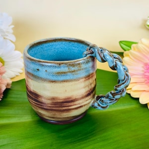 May include: A blue and brown ceramic mug with a unique rope-like handle. The mug has a light blue glaze with brown streaks and a light brown base.