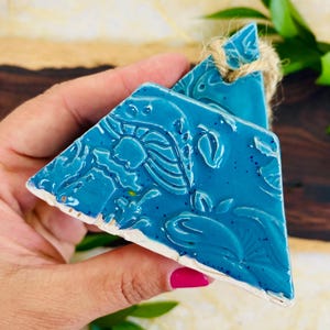 May include: A teal blue ceramic ornament with a floral design. The ornament is shaped like a triangle and has a rope loop for hanging.