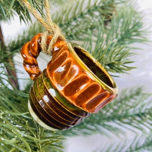 May include: A miniature ceramic mug ornament featuring a glossy brown and green glaze with a twisted handle. The mug is suspended by a piece of natural jute twine, showcasing a ribbed texture around the upper body and a smooth, rounded base.