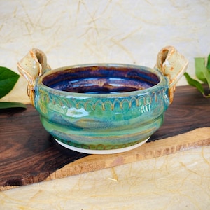 May include: A small, green and blue ceramic bowl with two handles. The bowl has a textured, wavy design and a white rim.