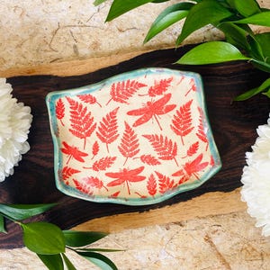 May include: A rectangular ceramic dish with a cream-colored background, adorned with red fern and dragonfly patterns. The dish has a light blue border and is set on a dark wooden surface, surrounded by white flowers and green foliage.