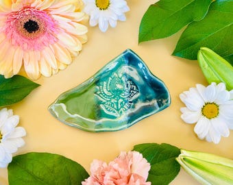May include: A teal green ceramic dish with a raised floral design. The dish is shaped like a leaf and has a glossy finish. It is surrounded by flowers and leaves.