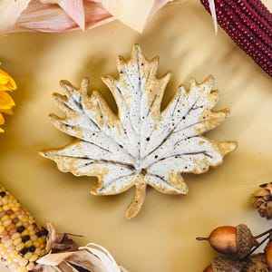 May include: A ceramic maple leaf-shaped dish with a white and speckled design, surrounded by fall harvest elements. The image includes a sunflower, corn, and acorns, creating a seasonal theme.