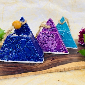 May include: Three ceramic triangular ornaments with a rope hanger and a wooden bead. The ornaments are decorated with a floral pattern and are painted in shades of blue, purple, and teal.