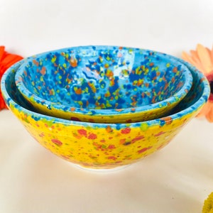 May include: Two ceramic bowls, one nested inside the other. The larger bowl is yellow with red and green speckles. The smaller bowl is blue with yellow and orange speckles.