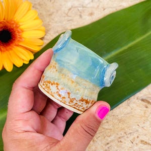 May include: A small, handmade ceramic cup with a unique design. The cup features a light blue and tan color scheme with brown speckles. The cup has two small handles and is held in a hand. A yellow flower and green leaf are in the background.