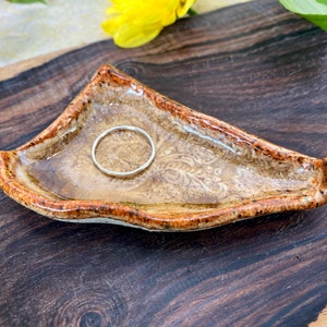 May include: A small, brown ceramic ring dish with a white and brown floral design. A silver ring is resting in the dish.