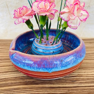 May include: A ceramic flower vase with a blue, red, and purple glaze holds pink and white carnations. The vase has a round shape with a smaller, perforated insert for arranging the flower stems. The flowers have pink edges.