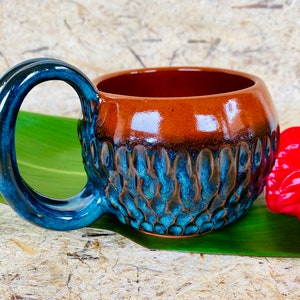 May include: A handmade ceramic mug with a brown and blue glaze. The mug has a unique textured design and a large, rounded handle.