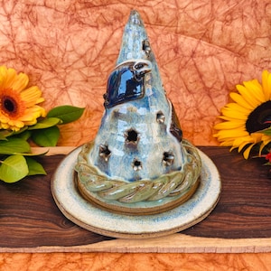May include: A handmade ceramic candle holder with a conical shape, glazed in shades of blue and brown. The holder features decorative cutouts and a braided green border, resting on a round plate. The candle holder is displayed on a wooden surface.