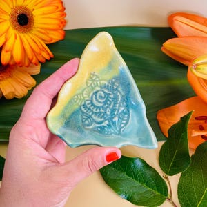 May include: A triangular ceramic dish with a yellow and green glaze. The dish has a floral design in the center.