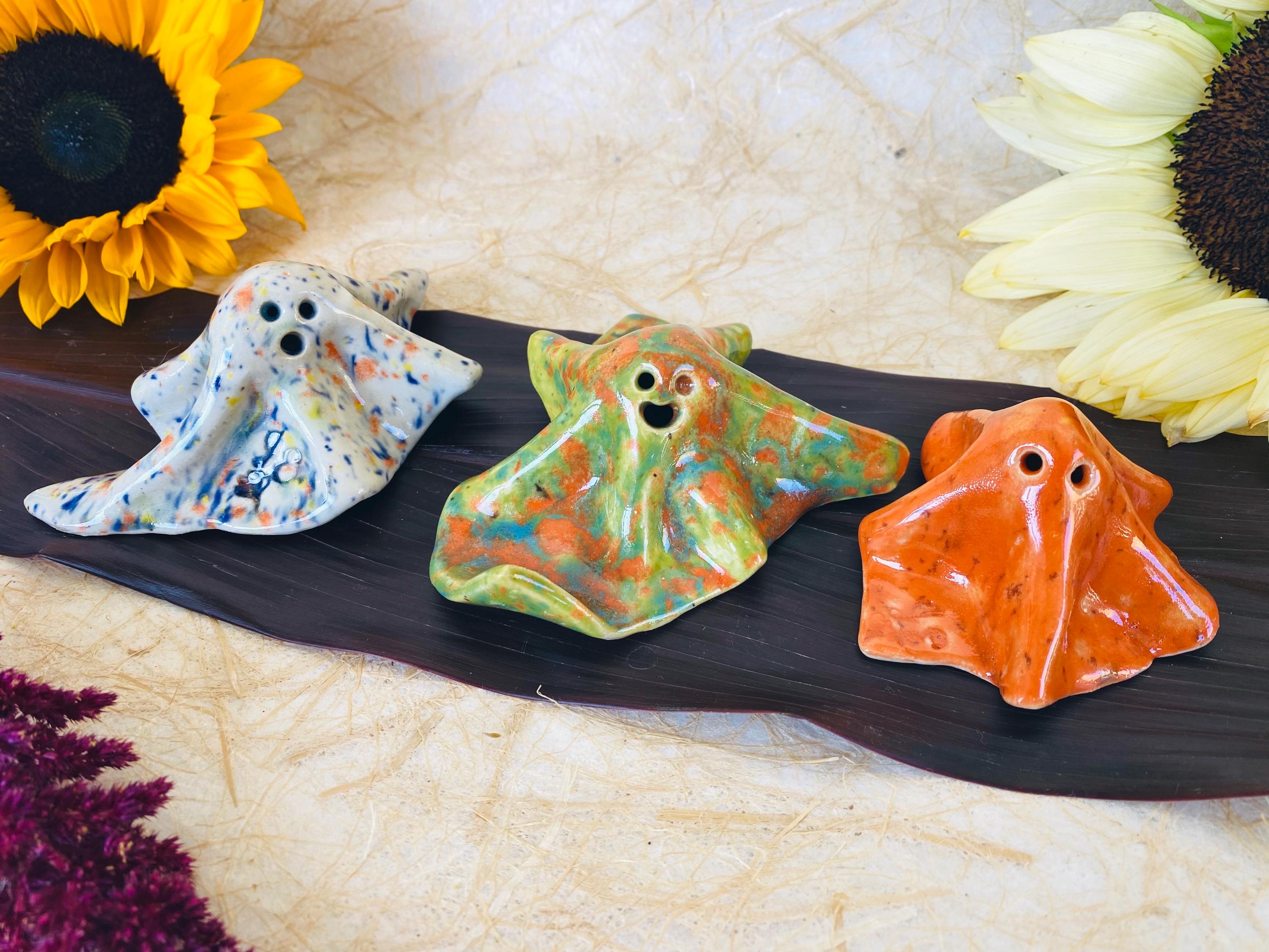 May include: Three ceramic ghost figurines with glossy finishes. The left ghost is white with multicolored speckles. The center ghost features a marbled blend of green and orange. The right ghost is a solid, vibrant orange with subtle dark flecks.
