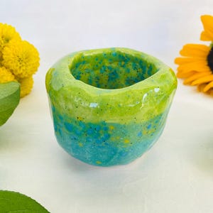 May include: A small, handmade ceramic planter with a green and blue glaze. The planter has a rough, textured surface and is perfect for displaying small plants or succulents.