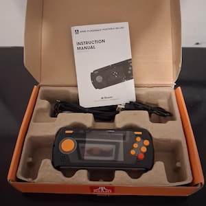 May include: An Atari Flashback Portable Deluxe handheld game console in a cardboard box with an instruction manual and a black charging cable. The console is black with orange buttons and a red button. The manual features the Atari logo and text.