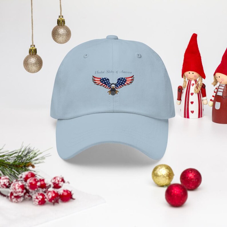Puede incluir: Gorra de b&eacute;isbol azul claro con un dise&ntilde;o bordado de un &aacute;guila con los colores de la bandera estadounidense. El texto "United States of America" est&aacute; encima del &aacute;guila. La gorra tiene un dise&ntilde;o cl&aacute;sico de seis paneles.