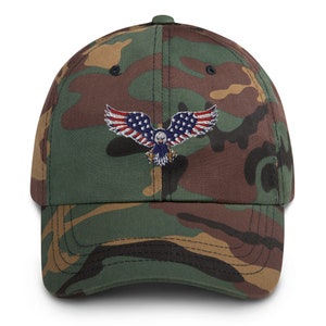 May include: A camouflage baseball cap with an embroidered eagle design. The eagle's wings are decorated with the stars and stripes of the American flag. The cap has a curved peak and a button on the crown.