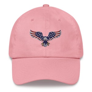 May include: A pale pink baseball cap with an embroidered eagle design. The eagle features the American flag on its wings. The cap has a curved peak and an adjustable strap at the back. The design is patriotic.