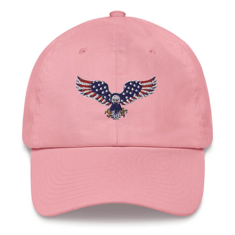 May include: A pale pink baseball cap with an embroidered eagle design. The eagle features the American flag on its wings. The cap has a curved peak and an adjustable strap at the back. The design is patriotic.