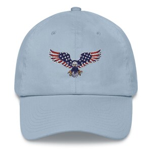 May include: A pale blue baseball cap with an embroidered eagle design. The eagle's wings are red and white striped, with a blue body and stars. The cap has a curved peak and an adjustable strap at the back.