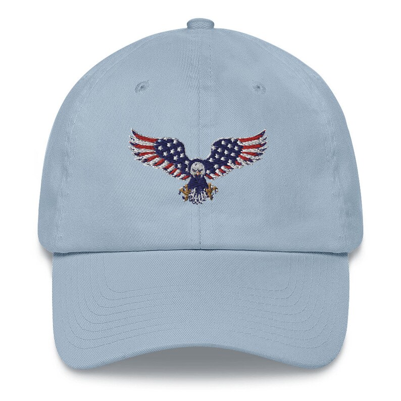 May include: A pale blue baseball cap with an embroidered eagle design. The eagle's wings are red and white striped, with a blue body and stars. The cap has a curved peak and an adjustable strap at the back.