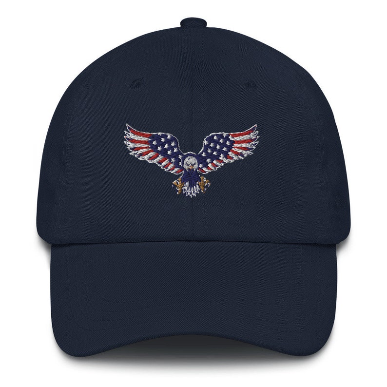 May include: A navy blue baseball cap with an embroidered eagle design. The eagle's wings are red and white striped, with a blue body and stars, resembling the American flag. A classic, adjustable hat.