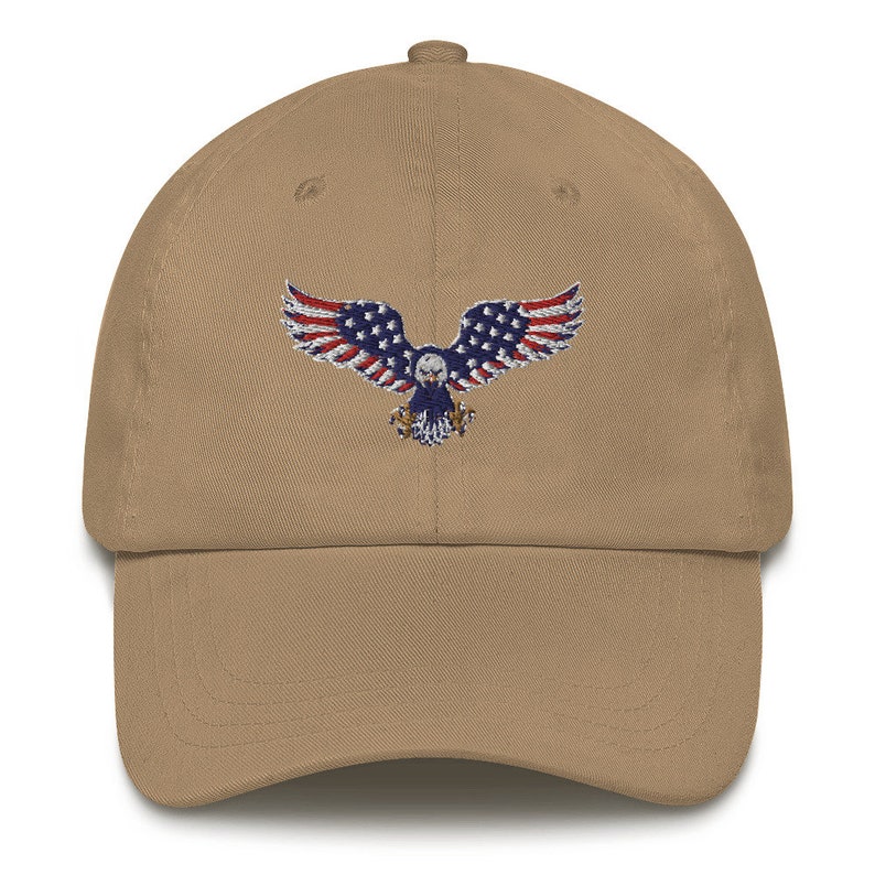 May include: A tan baseball cap with an embroidered design of an eagle with the American flag. The eagle's wings are red and white striped, and its body is blue with white stars. The cap has a curved peak and a fabric strap.