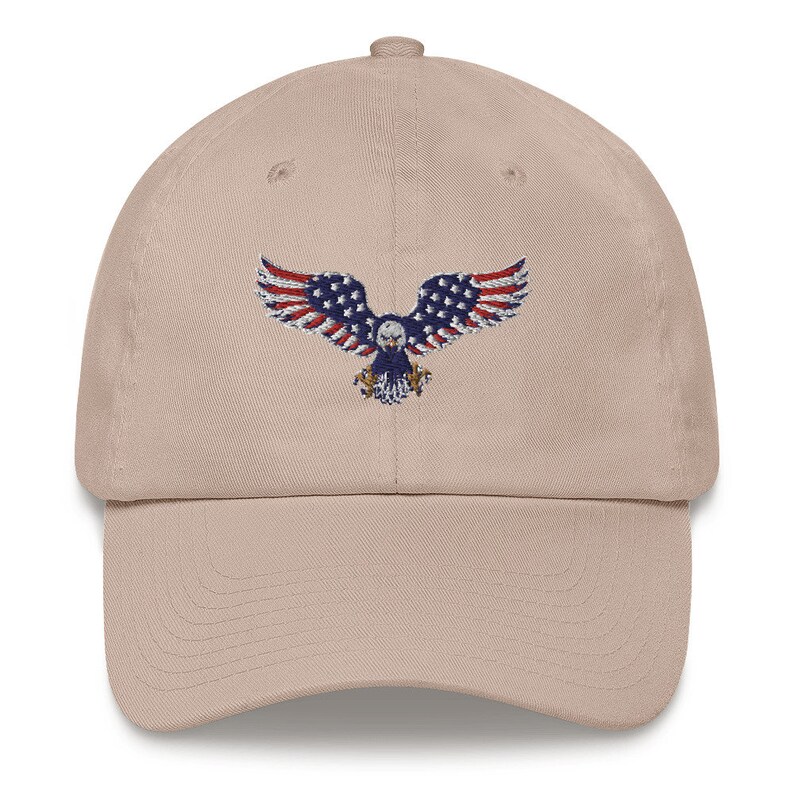 May include: A beige baseball cap with an embroidered eagle design. The eagle's wings are decorated with the stars and stripes of the American flag in red, white, and blue. The cap has a curved peak and an adjustable strap at the back.