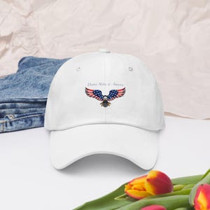 Puede incluir: Gorra de b&eacute;isbol blanca con un dise&ntilde;o de &aacute;guila americana. El dise&ntilde;o incorpora los colores de la bandera estadounidense: rojo, blanco y azul, con el texto "United States of America". La gorra est&aacute; hecha de un material suave.