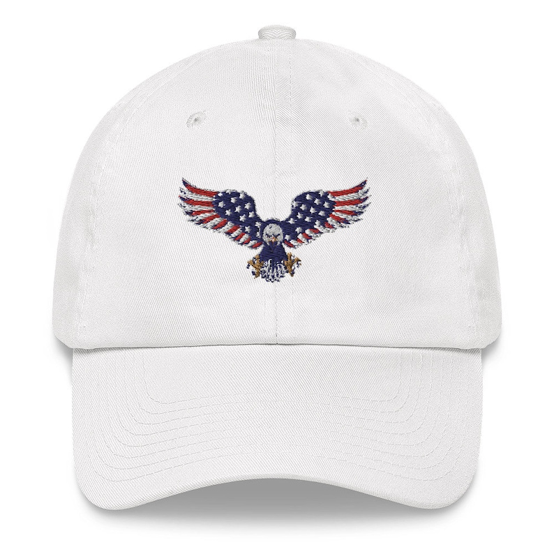May include: White baseball cap with an embroidered eagle design, incorporating the American flag. The eagle's wings are red and white striped, with a blue body and stars. A classic, adjustable hat.