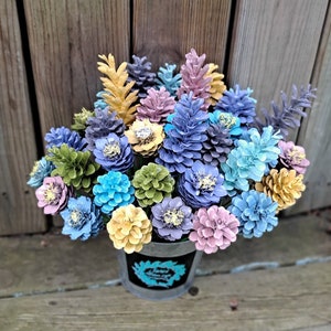 May include: A bouquet of painted pine cones arranged in a metal bucket. The pine cones are painted in shades of blue, green, yellow, and pink.