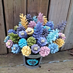 May include: A bouquet of painted pine cones in a variety of colours, including blue, pink, yellow, and green, arranged in a galvanised metal bucket. The pine cones are arranged in a circular pattern, with the tallest pine cones in the centre and the shorter pine cones around the edges.